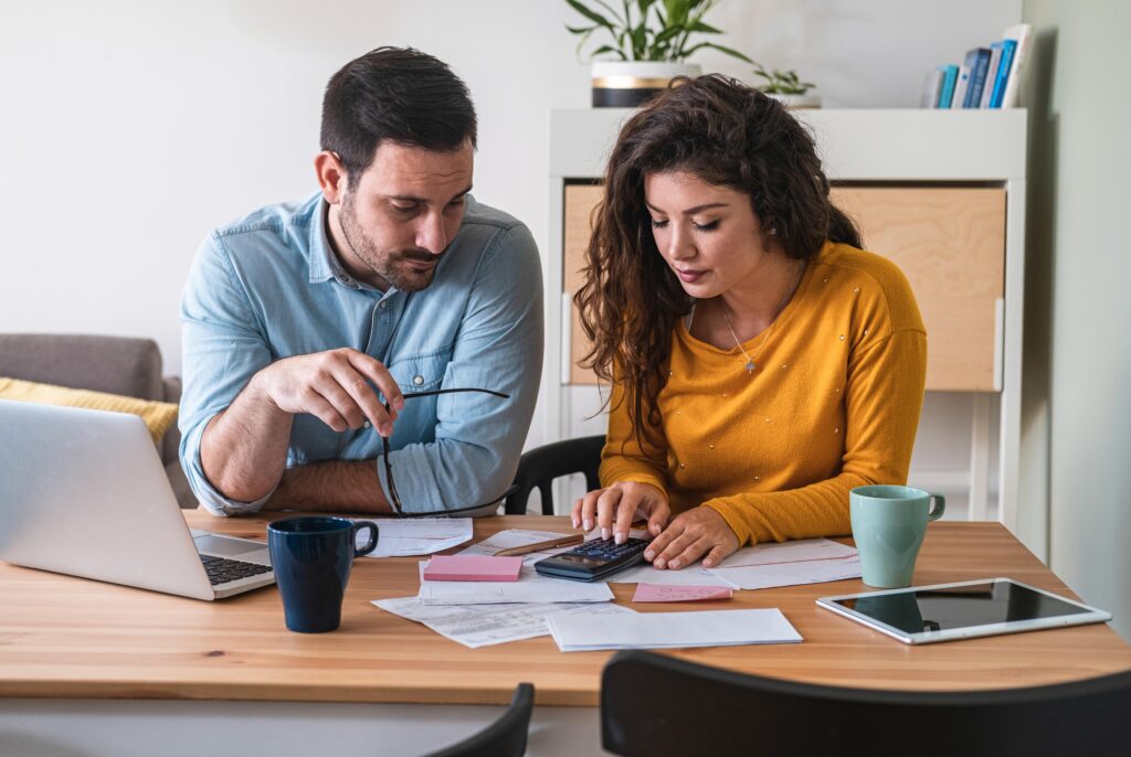Two coworkers sit at a wooden table reviewing documents with a laptop, calculator, and sticky notes nearby, in a bright home office environment.